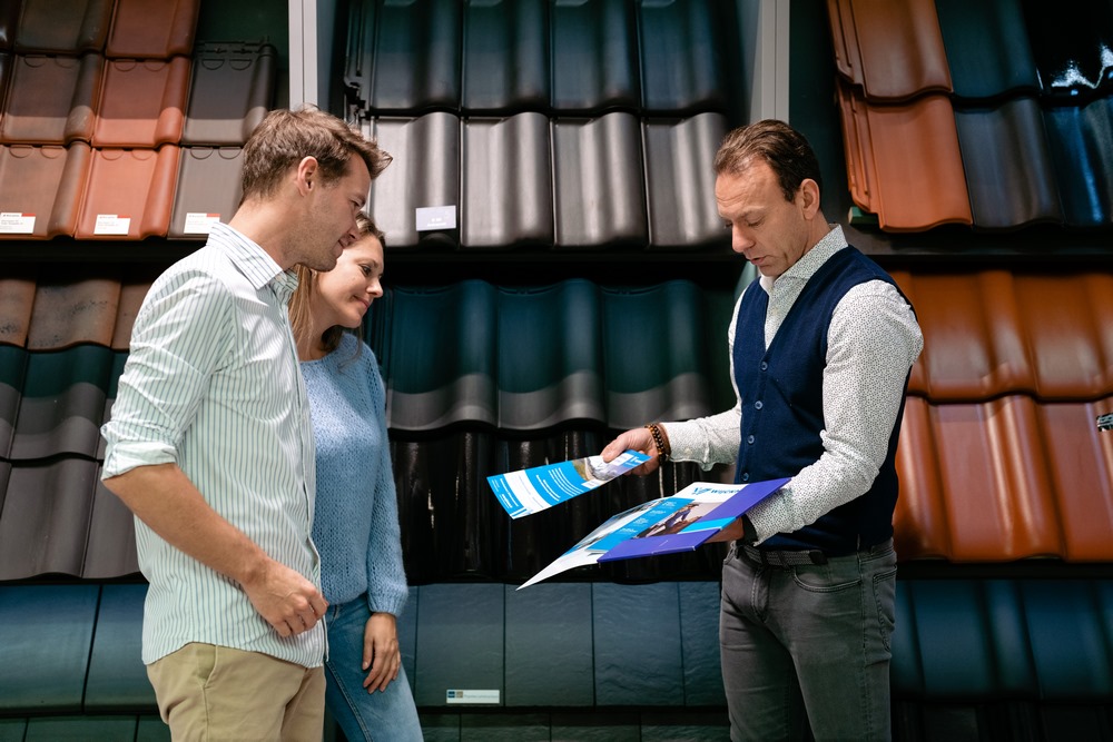 Verkoper geeft aan de hand van brochure info aan klanten over dakbedekking, met dakpannen op de achtergrond.